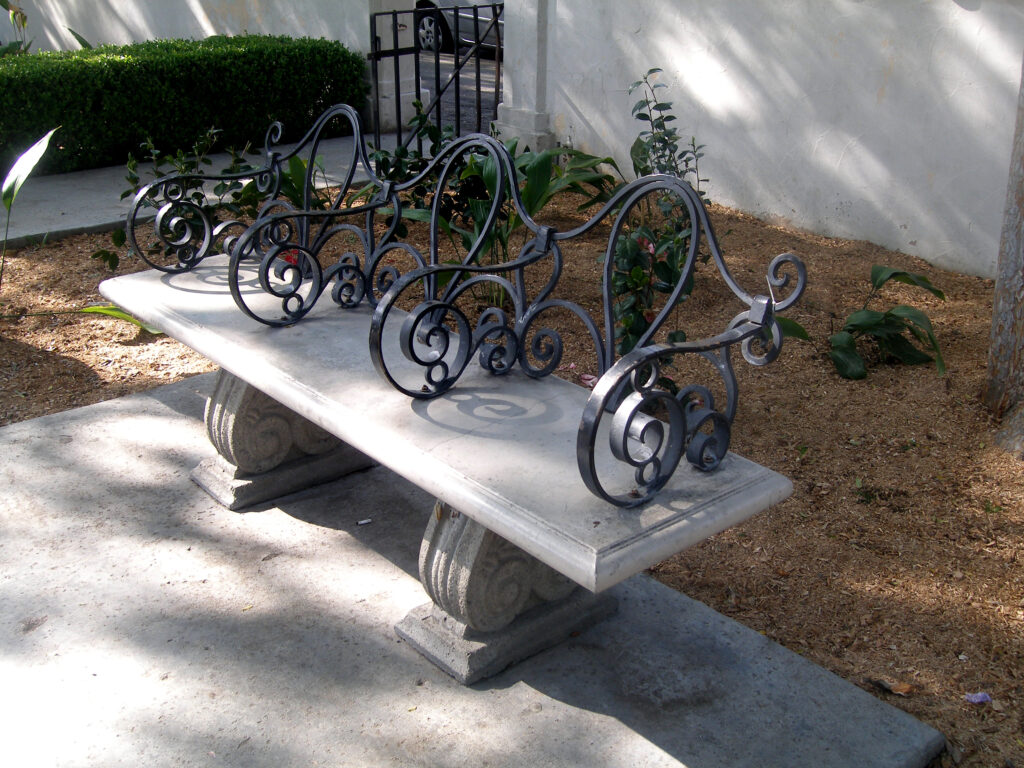 Image shows a concrete bench with iron armrests and back support dividing the bench into three sections, displaying a form a hostile architecture. 