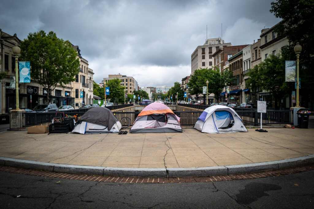 Three tents sit in the forefront of the image, on the sidewalk of a public space. The tents are in a line, sitting right before what appears to be a canal or tunnel. The image is clean, with few items littering the space.